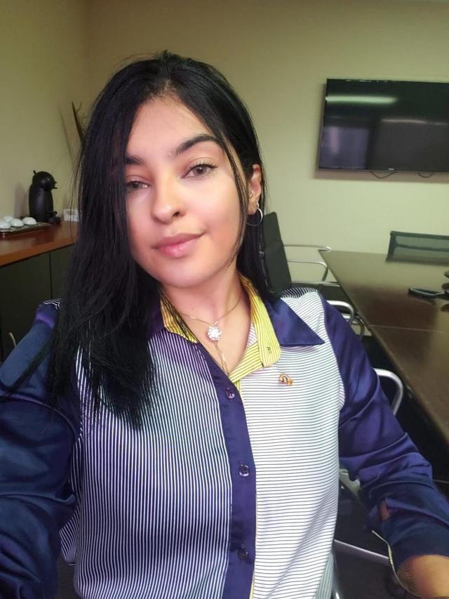 Foto da Michelle, uma mulher jovem e branca de cabelos pretos e olhos castanhos claros, usando brincos e colar, com uma camisa listrada com detalhes roxos e amarelos